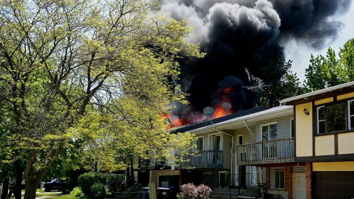 Dramatic house fire with thick smoke rising in a suburban neighborhood, surrounded by trees.