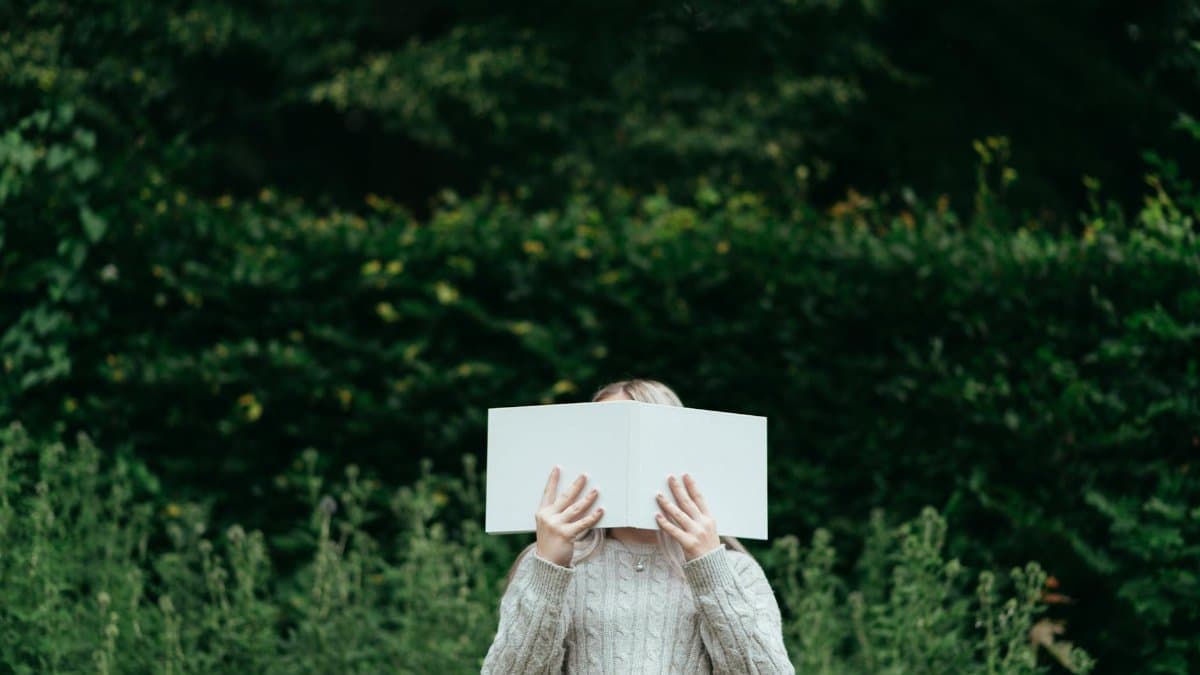 Unrecognizable female standing among green plants and hiding face with book while spending time in nature