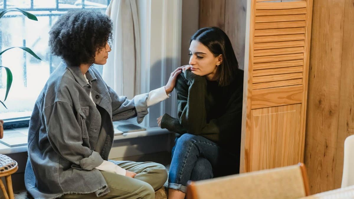 Calm young multiracial female best friends in casual clothes sitting near window and home and discussing discussing problems