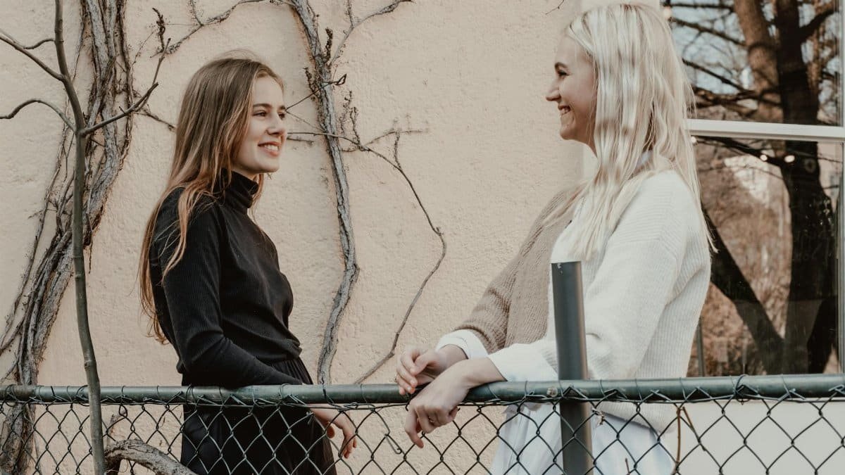 Two women having a cheerful conversation outdoors by a chain link fence on a sunny day.