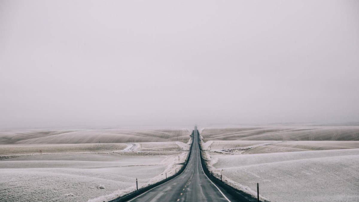 A long, empty road stretches through a wintery, snow-covered landscape under a cloudy sky.