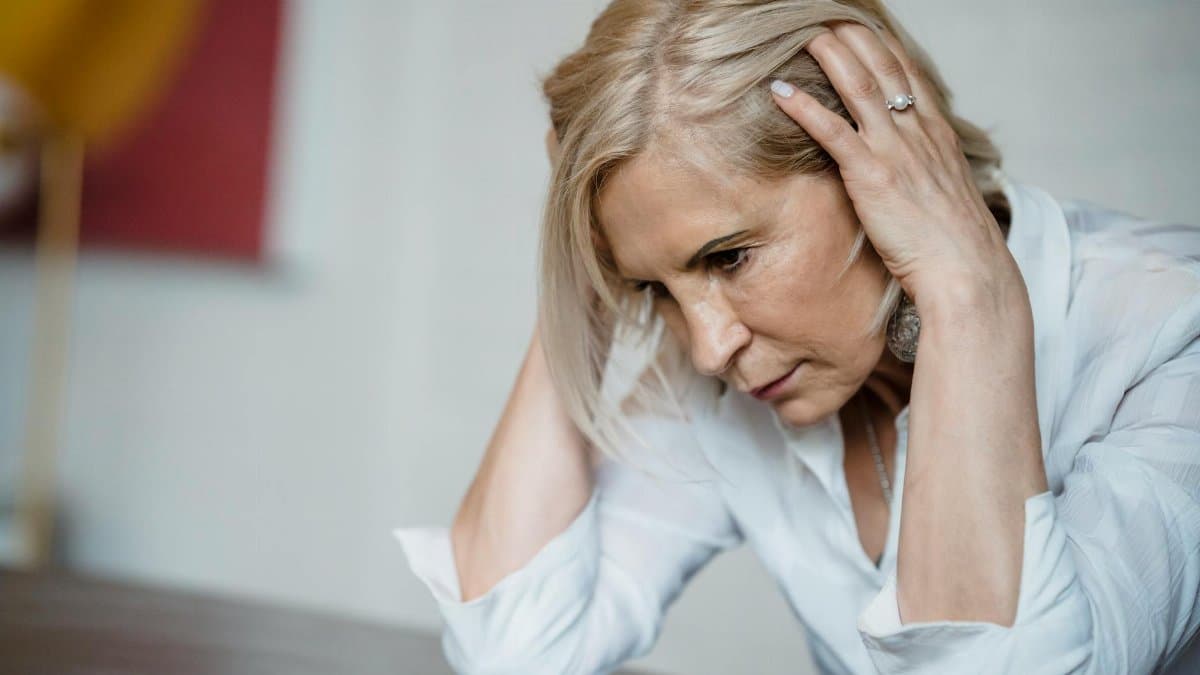 Woman with long blonde hair, deep in thought, wearing a white blouse indoors.
