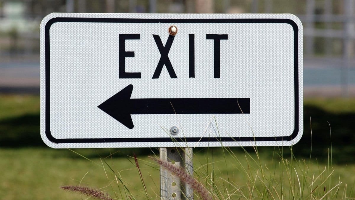 Exit sign with left-pointing arrow in a grassy outdoor setting.