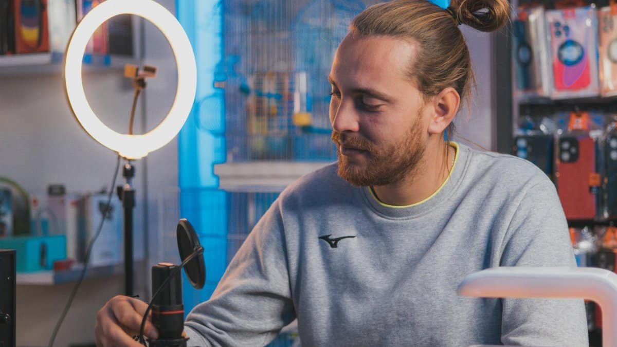 A man sitting in a studio setting adjusting a microphone with a ring light and equipment around.