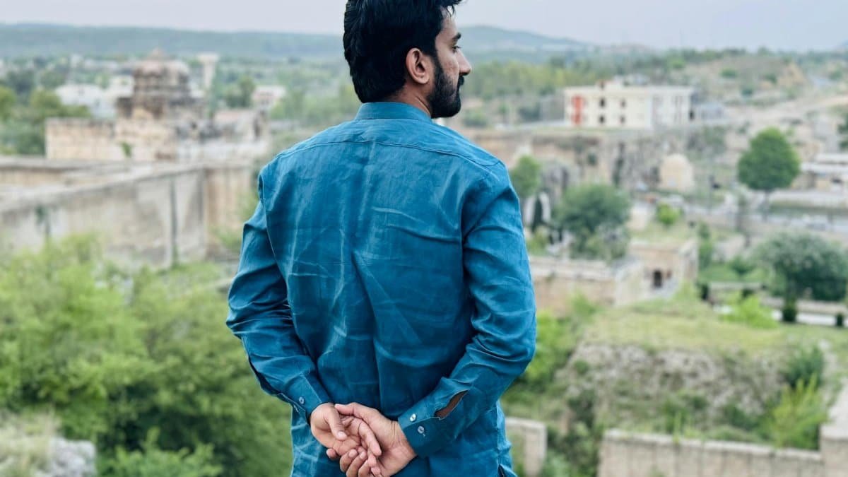 Man in blue attire stands with hands behind back, gazing at a serene outdoor landscape.