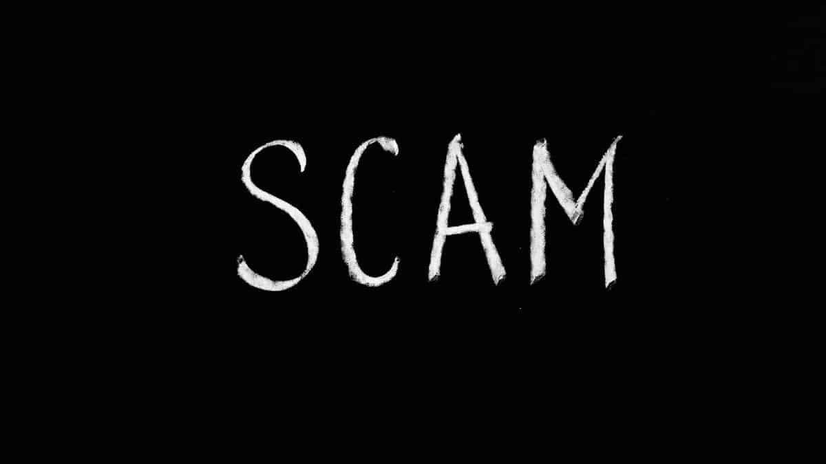 Chalk-drawn text 'SCAM' on black background, representing fraud and deception.