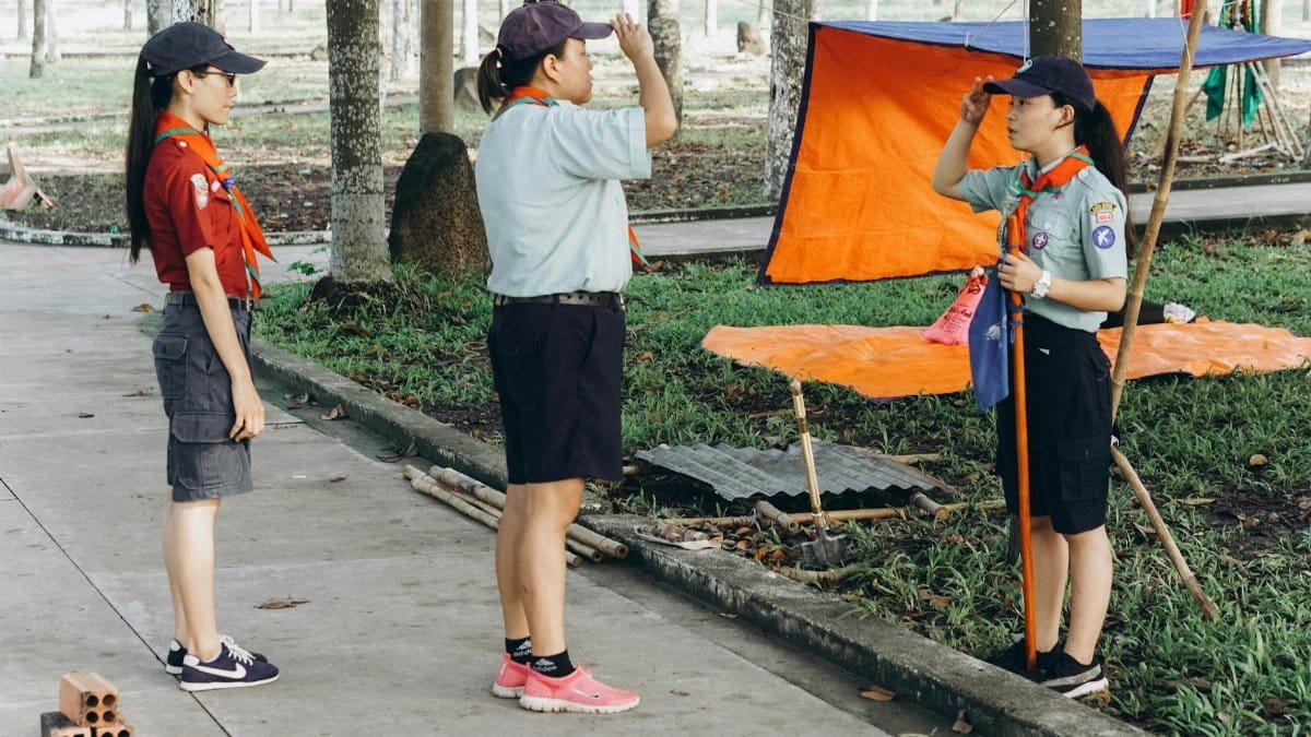 Three scouts salute in a park, participating in an outdoor scouting activity with orange tarp setup.
