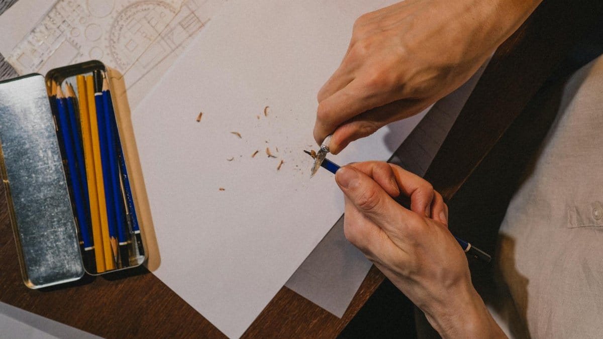 A close-up shot of a person sharpening a pencil over paper, surrounded by drawing tools indoors.
