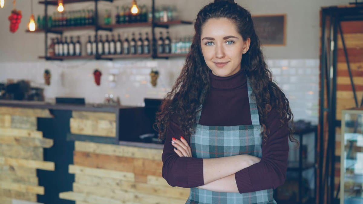 Confident café owner stands with arms crossed inside a rustic, cozy coffee shop.