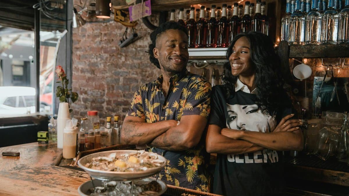 Two Black business owners smiling confidently behind a bar with oysters, embodying success and pride.