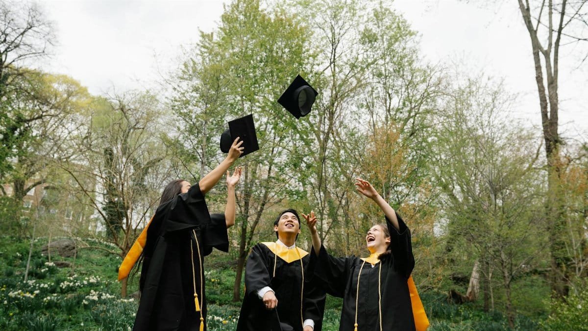 Three graduates celebrating and throwing caps in the air in a lush green park.