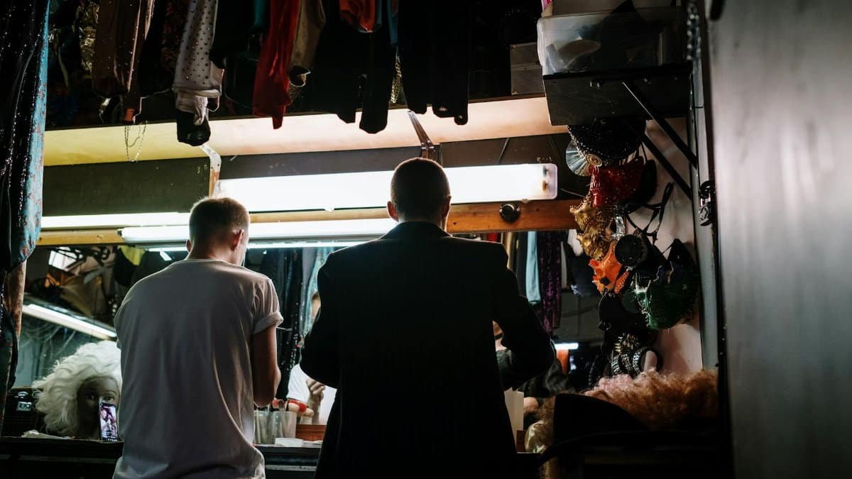 Two individuals preparing in a dimly lit dressing room with costumes and wigs, creating an artistic backstage ambiance.