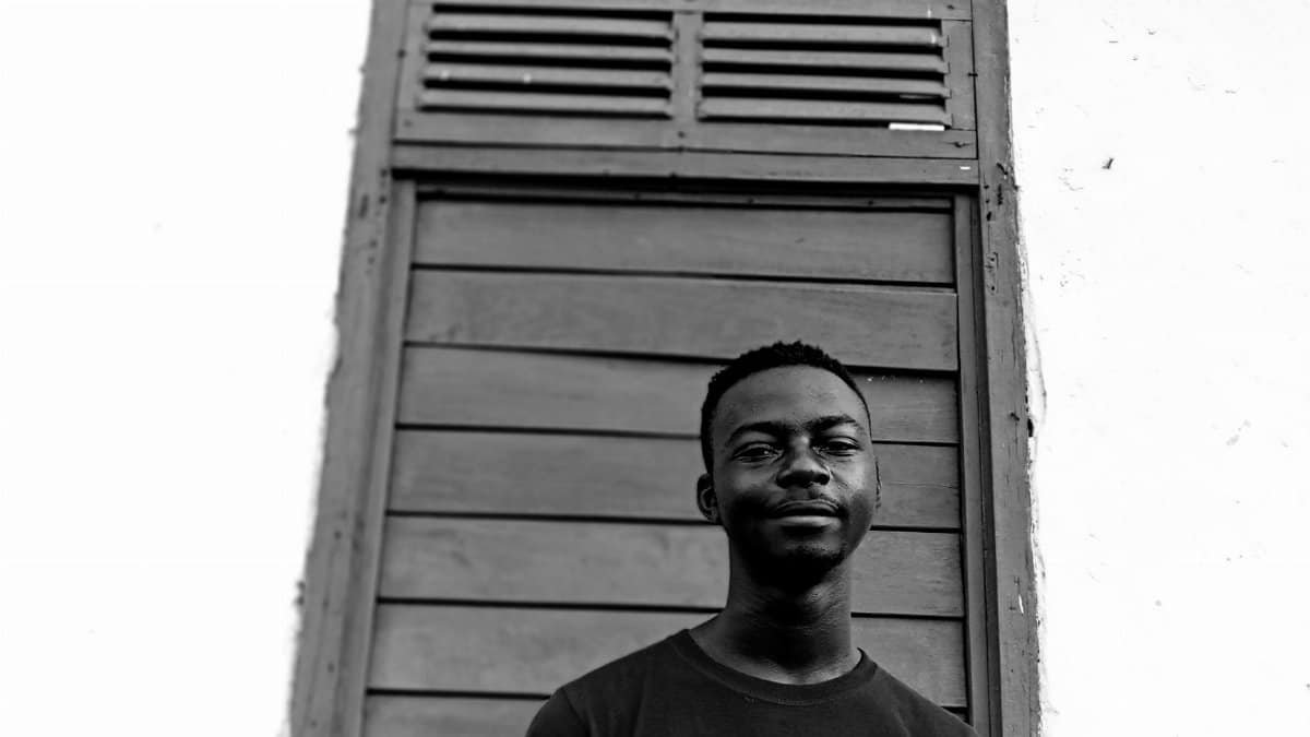 Black and white low angle of content African American male looking at camera while standing on street near shabby shutters