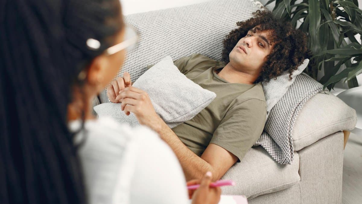 Man lying on couch during therapy session with female therapist taking notes.