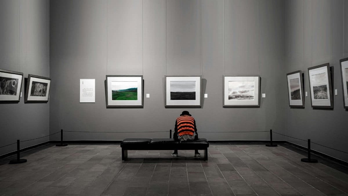 A solitary figure sitting in a contemporary art gallery surrounded by framed artworks.