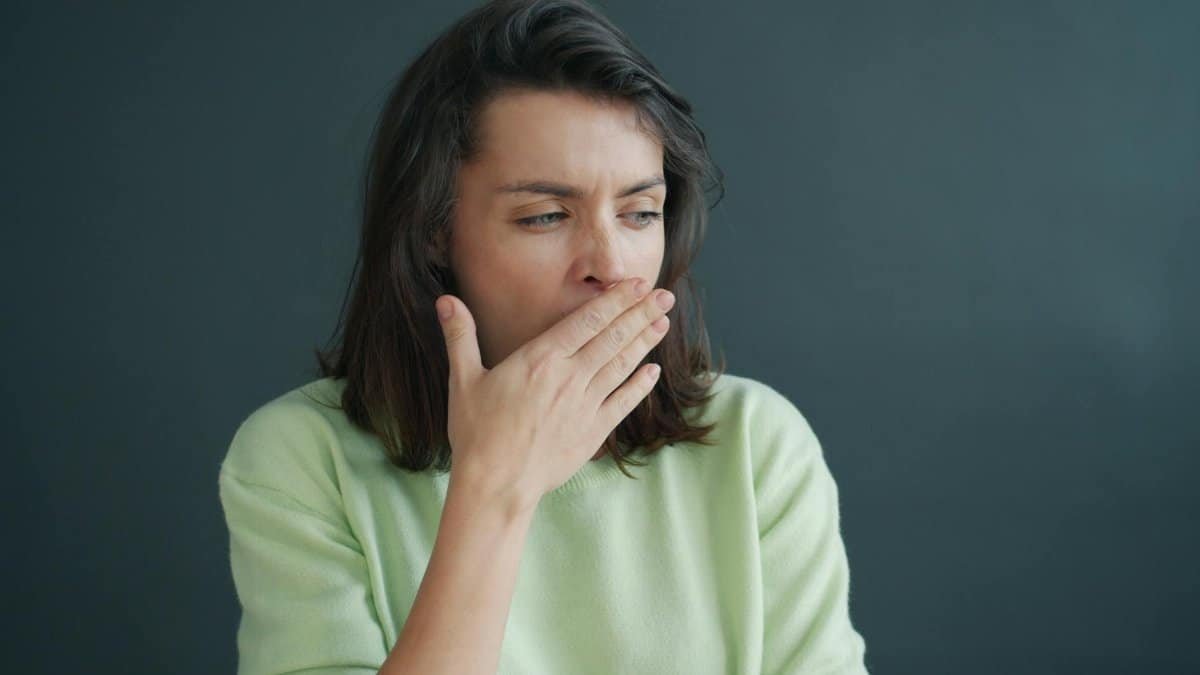 Young woman yawning, covering mouth with hand, wearing casual sweater against a gray background indoors.
