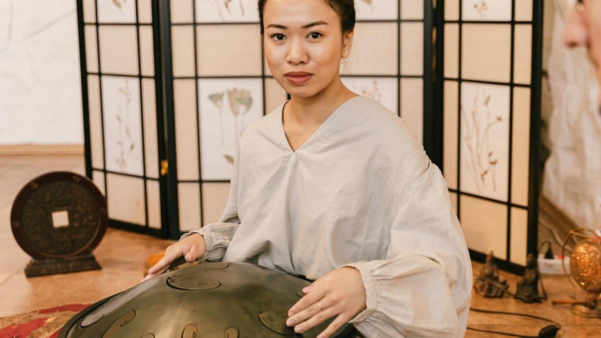 Asian woman playing handpan, creating healing music for relaxation in a peaceful indoor Zen setting.