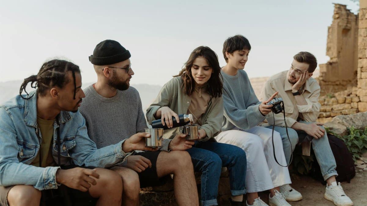 A diverse group of friends enjoy hiking together, sitting and sharing drinks outdoors on a sunny day.