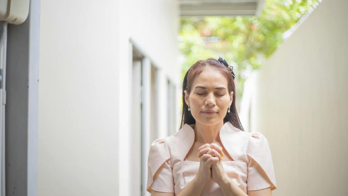 A serene woman with eyes closed, standing outdoors and clasping her hands in prayer, projecting calm and peace.