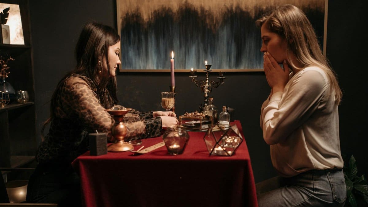 Two women engrossed in a mystical fortune telling session with candles and tarot cards on a dimly lit table.
