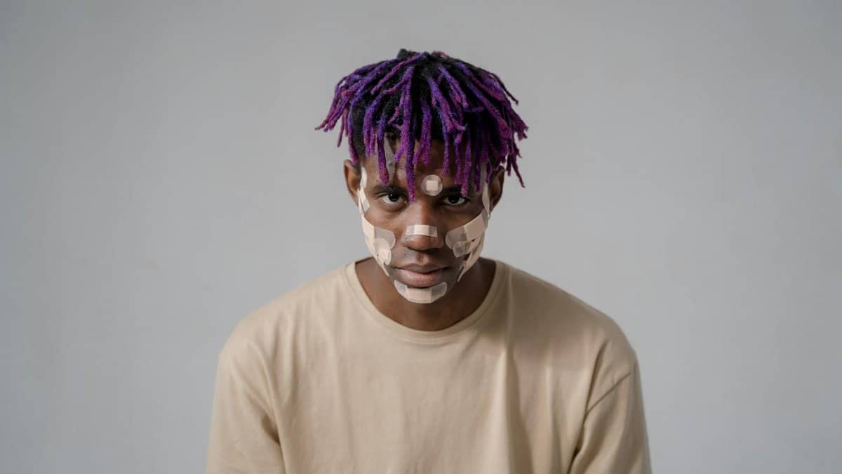 A portrait of a man with purple hair wearing adhesive bandages on his face, evoking themes of healing and self-expression.
