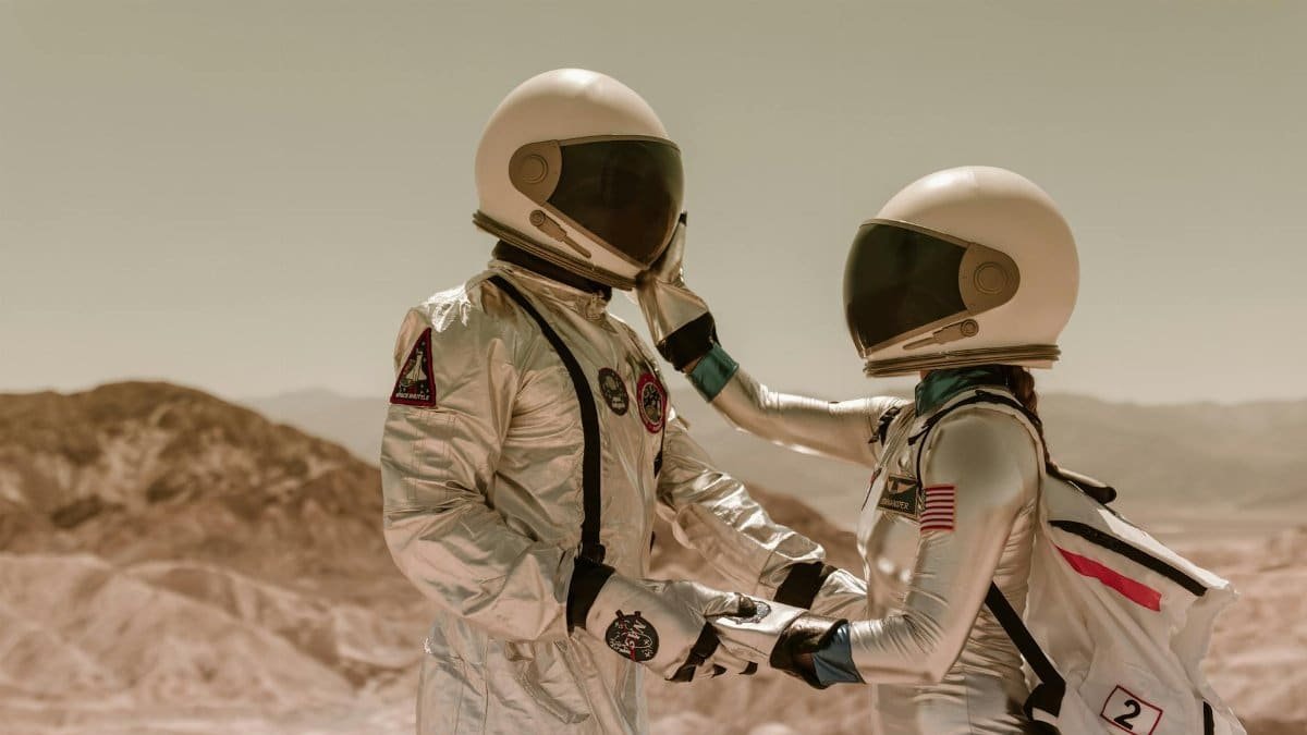 Two astronauts in space suits share a tender moment, face to face, in a desert setting.