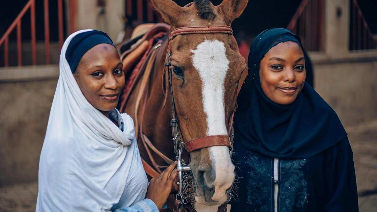 Two women dressed in traditional attire with a horse in a Nigerian stable setting. Cultural scene from Kano, Nigeria.