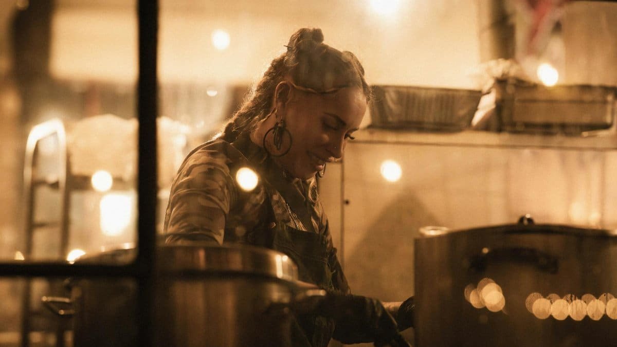An atmospheric view through a window of a female chef cooking at night in a warmly lit restaurant kitchen.