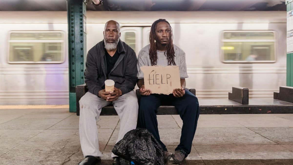 Two men sitting in a subway station with a cardboard sign reading 'HELP', highlighting social issues.