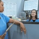Nurses in scrub suits discussing medical tasks in a clinical environment.