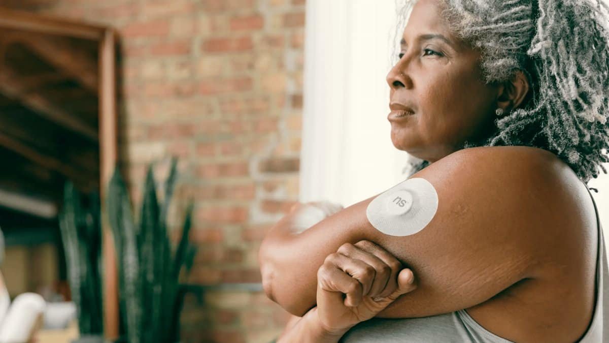 A mature woman with gray hair wearing a continuous glucose monitoring device on her arm.