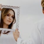 A woman posing with an ornate frame held by a man, outdoors in daylight.
