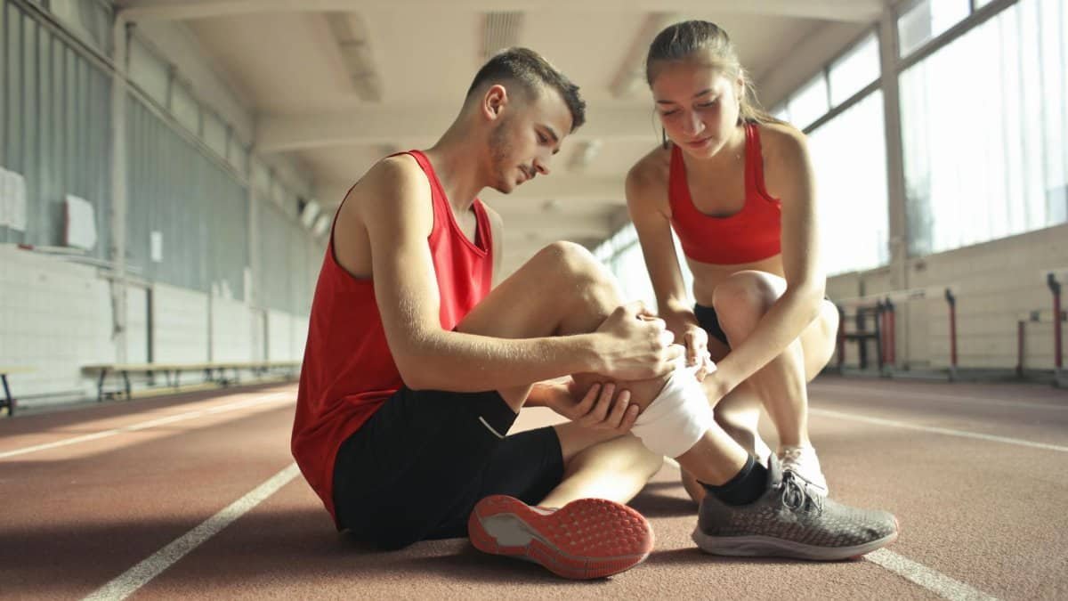 Side view of athlete sitting on rubber track in sports hall while sportive female partner helping to band leg after injury