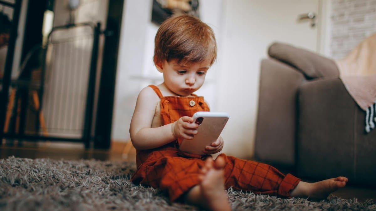 A young child wearing orange overalls is captivated by a smartphone indoors.