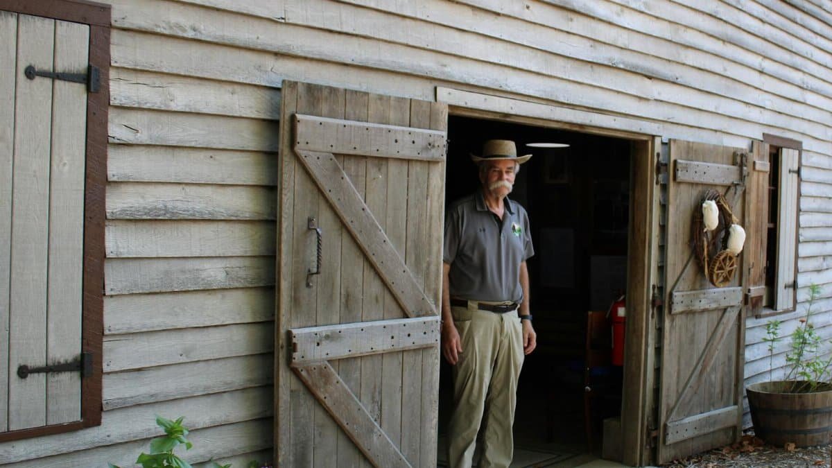 Man standing in wooden barn doorway wearing a hat, showcasing a rustic setting.