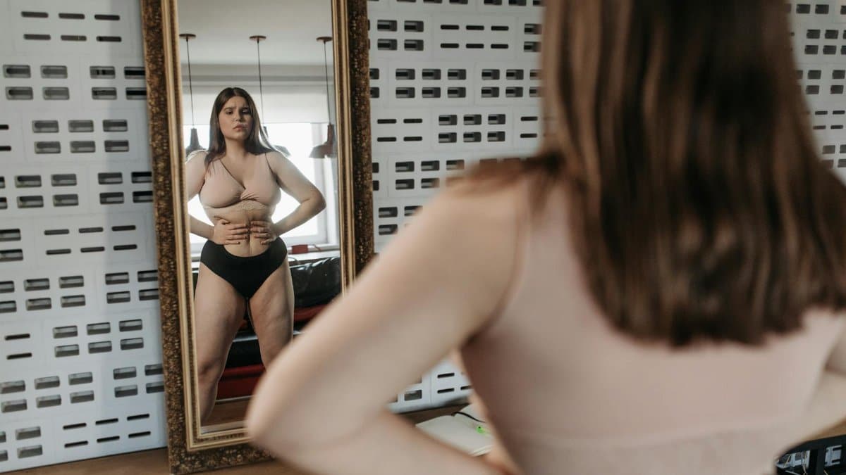 Woman in underwear looking at her reflection in a mirror, contemplating body image.
