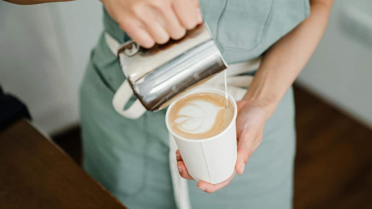 High angle of crop anonymous young barista in apron pouring milk into disposable cup and making latte art