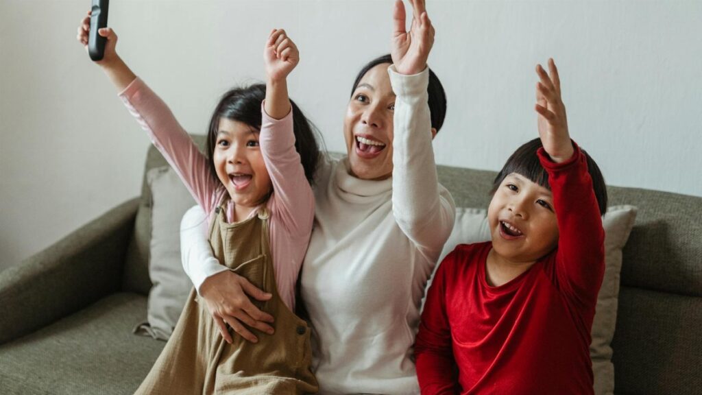 Cheerful Asian mom and kids watching sport on TV at home and screaming happily while cheering with raised arms