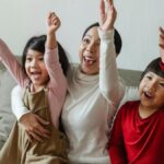 Cheerful Asian mom and kids watching sport on TV at home and screaming happily while cheering with raised arms