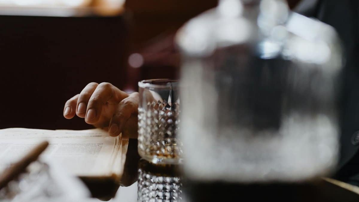 A hand casually rests on a book beside a crystalline whiskey glass in a warm indoor setting.