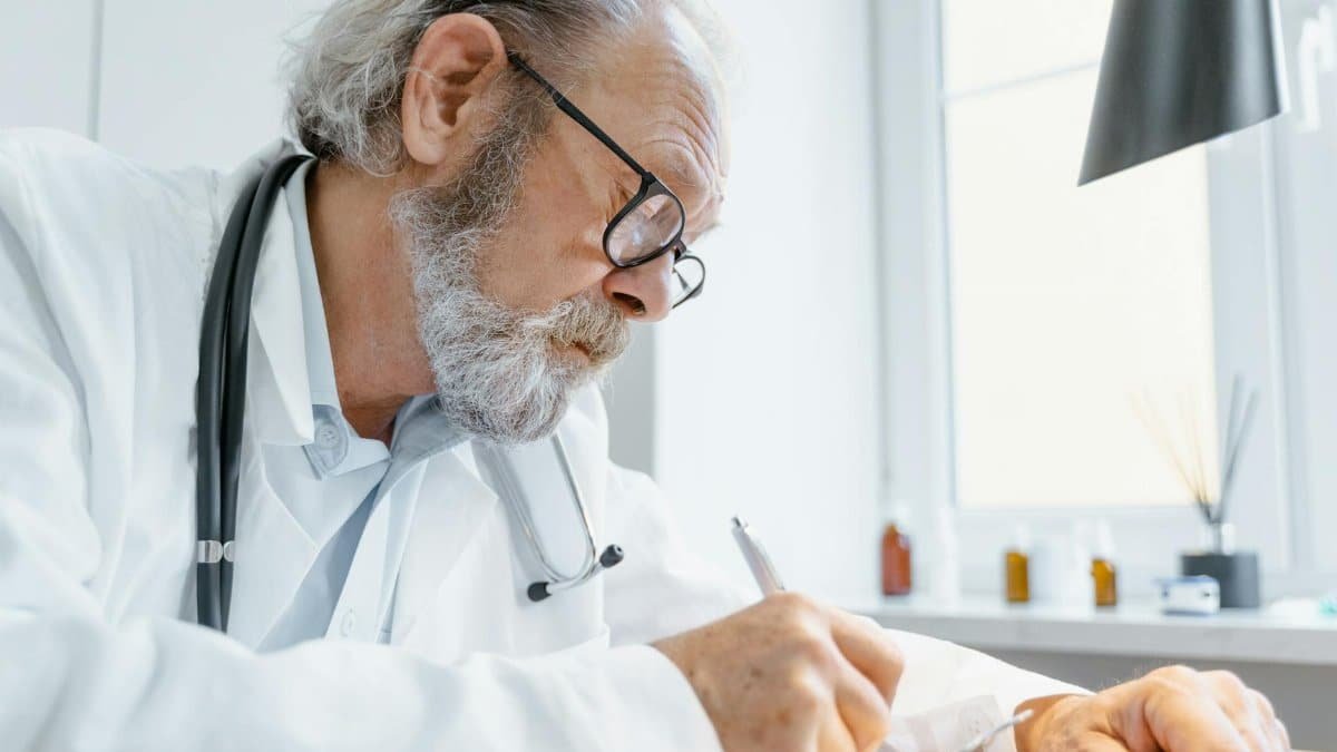 Elderly male doctor writing notes in a bright medical office.