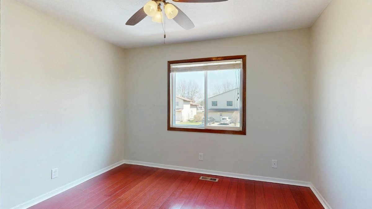 Spacious empty room with wooden flooring, window view, and ceiling fan.