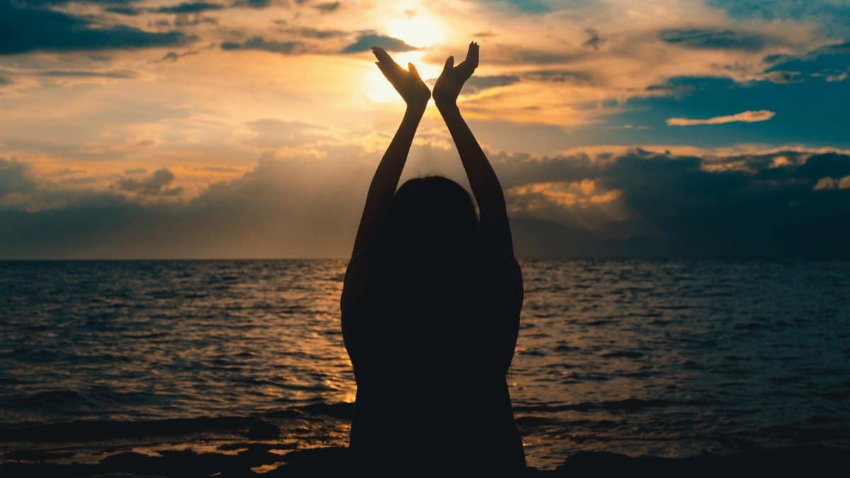 A silhouetted figure raises hands towards the setting sun over a serene ocean view.