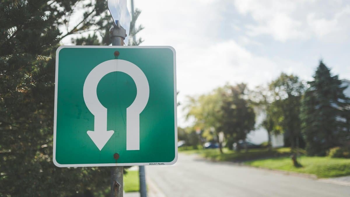 Close-up of a green U-turn road sign on a sunny suburban street with trees.