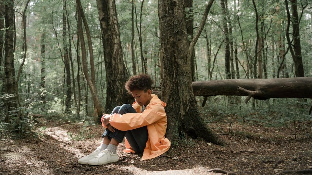 A young boy in a bright jacket sits alone in a serene forest, evoking emotions of solitude and reflection.