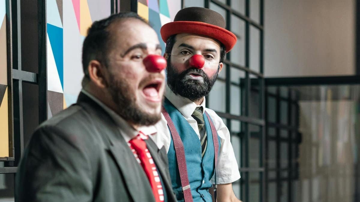 Two male clowns with red noses perform expressively on stage, adding humor and charm.