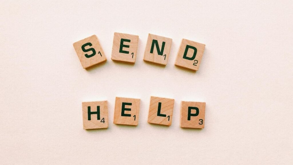 Wooden Scrabble tiles arranged to form the phrase 'send help' on a light pink background.
