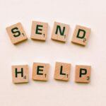 Wooden Scrabble tiles arranged to form the phrase 'send help' on a light pink background.