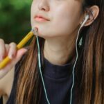 Crop young Asian lady touching chin with pencil while listening to music with earphones and dreaming on blurred background