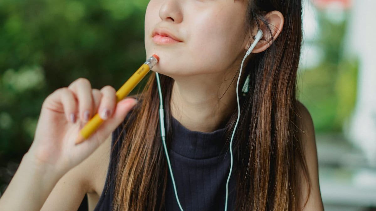 Crop young Asian lady touching chin with pencil while listening to music with earphones and dreaming on blurred background